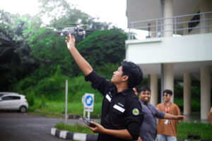 Pelatihan Drone Multispektral di kampus IPB, seorang pria berseragam hitam mengoperasikan drone dengan peserta yang antusias di latar belakang
